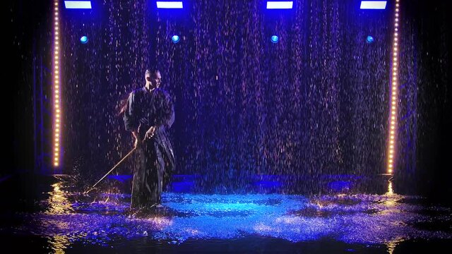 A male Japanese kendo fighter dressed in traditional kimono practices a martial art with a bamboo bokken in the rain. Slow motion.