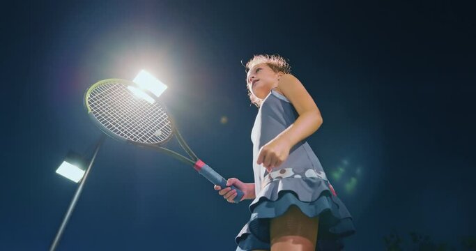 Teenage girl excited tennis win. Slow motion young female sportsman celebrates won match in tennis game. Low angle athlete girl with tennis racket making hand movement. Late night sport game 
