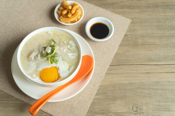 Top up view of rice porridge or congee with minced pork, egg, slice ginger and slice scallion topping near by deep-fried dough stick and soy sauce in a white bowl on wooden desk . Breakfast concept.