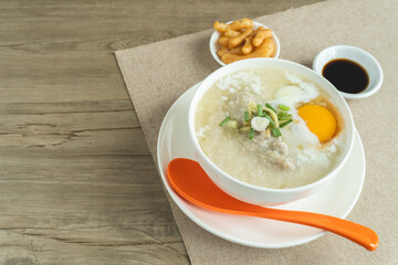 Rice porridge or congee with minced pork, egg, slice ginger and slice scallion topping near by deep-fried dough stick and soy sauce in a white bowl on wooden desk. Breakfast concept.