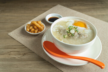 Rice porridge or congee with minced pork, egg, slice ginger and slice scallion topping near by deep-fried dough stick and soy sauce in a white bowl on wooden desk. Breakfast concept.