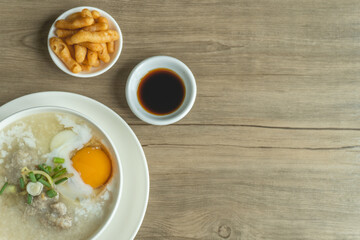 Close up view of rice porridge or congee with minced pork, egg, slice ginger and slice scallion topping near by deep-fried dough stick and soy sauce in a white bowl on wooden desk. Breakfast concept.