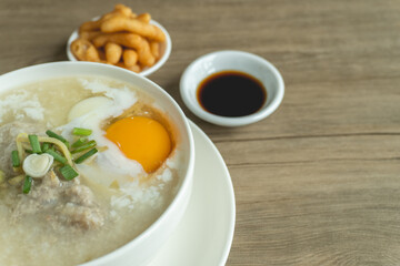 Close up view of rice porridge or congee with minced pork, egg, slice ginger and slice scallion topping near by deep-fried dough stick and soy sauce in a white bowl on wooden desk. Breakfast concept.