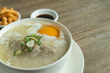 Close up view of rice porridge or congee with minced pork, egg, slice ginger and slice scallion topping near by deep-fried dough stick and soy sauce in a white bowl on wooden desk. Breakfast concept.