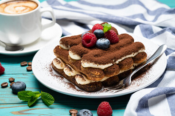 portion of Classic tiramisu dessert with raspberries and blueberries and cup of espresso coffee on blue wooden background
