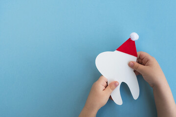 Little child holding cute decorative tooth in a red Santa Claus hat on light blue background....