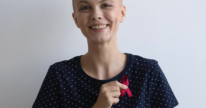On gray studio background standing hairless bald woman cancer patient with red awareness symbolic ribbon smile looking at camera. Donation and charity, supporting people with incurable disease concept