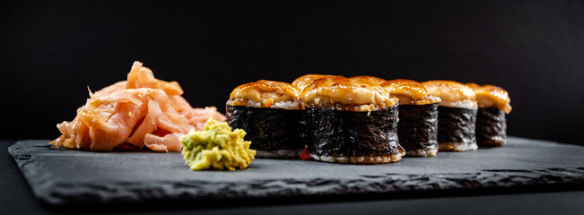 Panoramic photo of served warm sushi rolled in nori sheets with ginger and wasabi on a black stone plate.