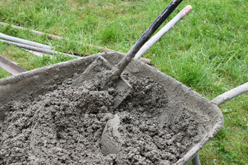 Wheelbarrow full of concrete mortar and shovel for building on green grass background outdoors.
