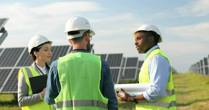 Three workers in special uniform discussing working moments outside. One woman and two men are talking on solar plantation. Teamwork.