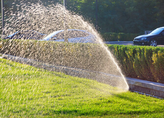 A beautiful spray fountain from an automatic sprinkler system sprays the green lawn on a bright...