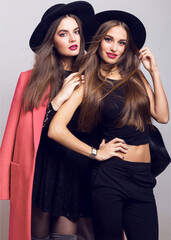 Fashion image of two girls, sisters, posing on grey background, hugging, smiling. Wearing stylish pink  coat and black hat, short top and evening dress.