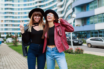 Fall fashion look.  Couple of attractive graceful girls in cute round glasses and black hats posing on background of business center.