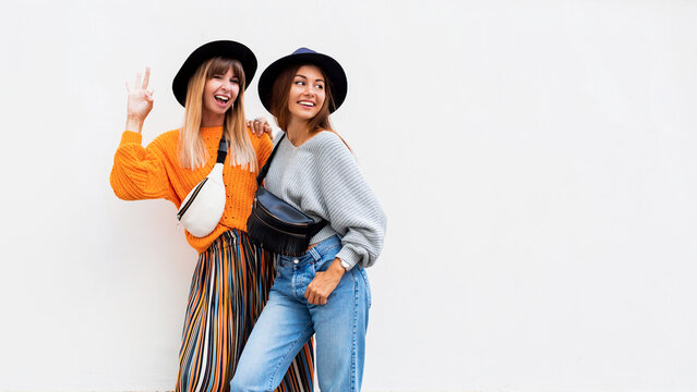 Best friends, couple of  stylish girls posing on white background. Fall season. Wearing stylish orange knitted sweater, black hat , bum bag. Friends spending great time together . Copy space .