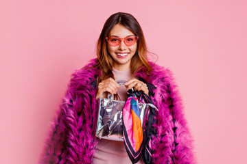 Wonderful girl in fancy pink faux fur short coat holding silver hand bag and looking forward.  Fashion studio shot. Winter season. Shopping concept.