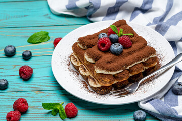 portion of Classic tiramisu dessert with raspberries and blueberries on blue wooden background
