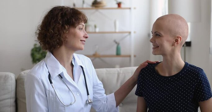 Home visiting cancer patient concept. Female oncologist doctor in white coat talks to bald girl successful recovered oncology disease remission, warm conversation and professional nursing care concept