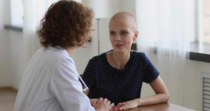 Optimistic cancer patient talks to oncologist doctor during home visit or meeting in clinic. Kind nurse hold hand of bald unhealthy female express support telling about future rehabilitation concept