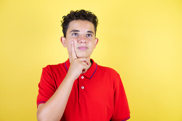 Beautiful teenager boy over isolated yellow background thinking with hand on chin and looking to...