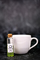 Small vial with cork and green liquid and poison skull label next to tea cup on dark background