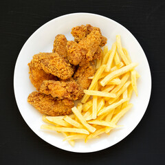 Homemade Crispy Chicken Wings and French Fries on a white plate on a black surface, top view. From above, overhead, flat lay.