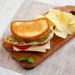 Homemade Pesto Chicken Sandwich with Potato Chips on a rustic wooden board on a white wooden background, side view. Close-up.
