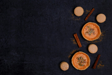 autumn orange creamy pumpkin pies for halloween with seeds, cinnamon sticks, several macaroon cakes on a black background, top view, place for text, flat lay