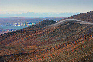 Autumn Arctic landscape. View of the mountain slope, tundra and the city of Anadyr in the distance. Fog on the mountain tops. Travel and Hiking in the Far North of Russia. Golden ridge, Chukotka.