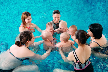 A group of mothers with their young children in a children's swimming class with a coach.