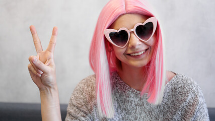 Portrait of a happy cheerful girl showing peace gesture. Woman wearing heart-shaped glasses and pink wig - positive portrait