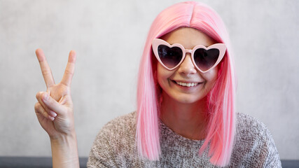 Portrait of a happy cheerful girl showing peace gesture. Woman wearing heart-shaped glasses and pink wig - positive portrait