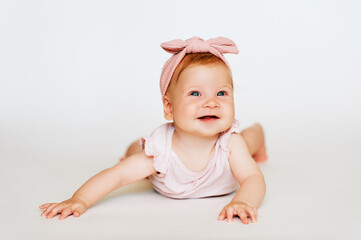 Portrait of adorable red-haired baby lying on belly, white background, wearing pink body and headband, looking up with smile