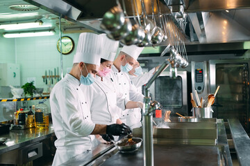 Chefs in protective masks and gloves prepare food in the kitchen of a restaurant or hotel.