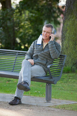 Mature man with face mask hanging on a ear talking on smart phone while sitting on a park bench