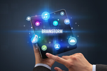 Naklejka premium Businessman holding a foldable smartphone with BRAINSTORM inscription, social media concept