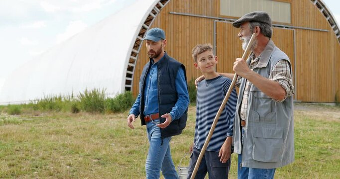 Caucasian young man with old father and small teen son walking outdoors at village shed and talking. Three male generations. Boy with dad and grandfather. Farming lifestyle. Countryside.