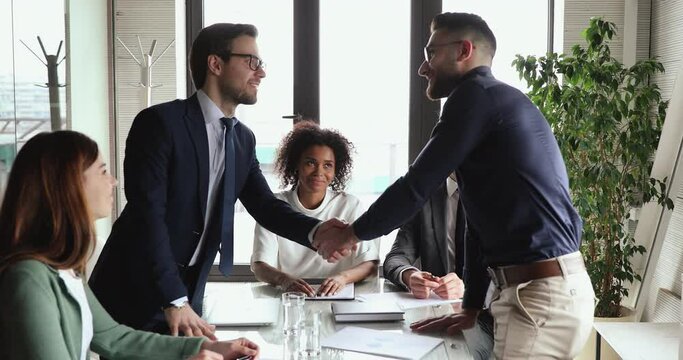Happy young businessman getting up from table to shake hands with arabian skilled expert, thanking for company financial situation or marketing strategy explaining, business agreement concept.
