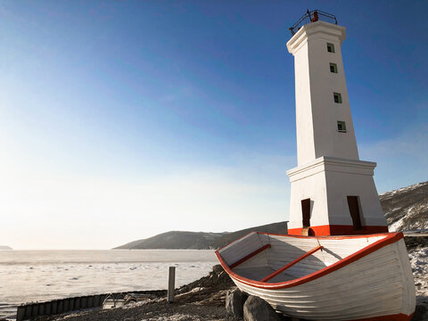 Picture of old lighthouse and wooden boat on the seashore of Magadan, Russia. Tall white building and boat with red stripes. Clear blue sky and ice on the sea in winter. Copy space for text