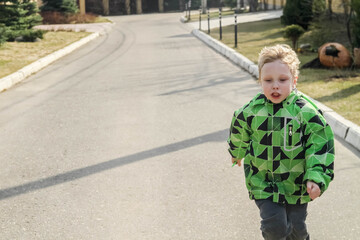 A blond European boy in a green jacket runs along the road.