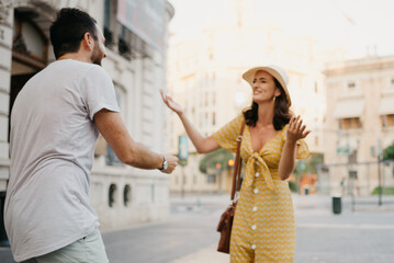 Fototapeta premium A laughing girl in a hat and a yellow dress with a plunging neckline is joking with her boyfriend with a beard and sunglasses in the old town. A couple of tourists on the sunset in Valencia.
