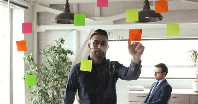 Focused young arab male manager putting colorful sticky paper notes on glass wall, managing working process in modern office. Skilled arabic scrum master in eyeglasses checking workflow indoors.