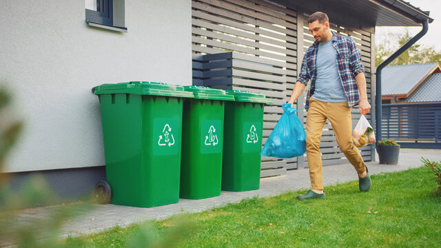 Caucasian Man Is Walking Outside His House In Order To Take Out Two Plastic Bags Of Trash. One Garbage Bag Is Sorted As Biological Food Waste, Other Is Recyclable Bottles Garbage Bin.