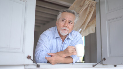 Fototapeta premium Senior asian man looking by window, holding cup of tea