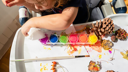 Toddler boy is home schooled paiting yellow leaves and pine cones in fall with eco friendly paint. Sensorial activity at home.