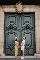 A girl in a hat and a dress with a plunging neckline and her boyfriend with a beard are staying near the giant doors holding each other's hand in the old town. A couple of tourists in Valencia.