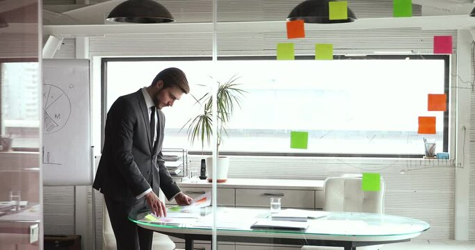 Concentrated motivates young successful businessman in formal suit attaching sticky notes on paper document or setting reminders on important report, involved in project management process in office.