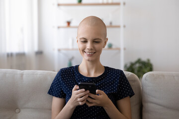 Happy millennial hairless Caucasian sick woman patient sit on sofa at home use modern cellphone...
