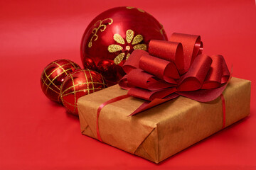 A gift in craft paper with a red bow lies next to New Year's red-gold balls on a red background, close-up