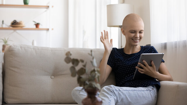 Smiling hairless ill female suffering from cancer relax at home talk on video call on tablet. Happy bald sick young woman struggling with oncology have webcam virtual talk conference on pad gadget.