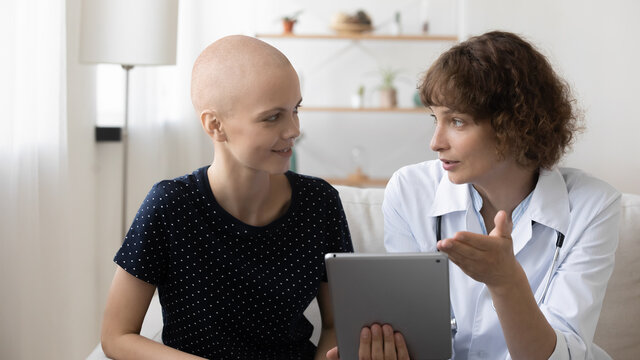 Doctor And Young Hairless Sick Female Cancer Patient Look At Tablet Screen Discuss Treatment Or Cure Together. Caring Nurse Talk With Ill Bald Woman Suffer From Oncology, Consider Good Results On Pad.